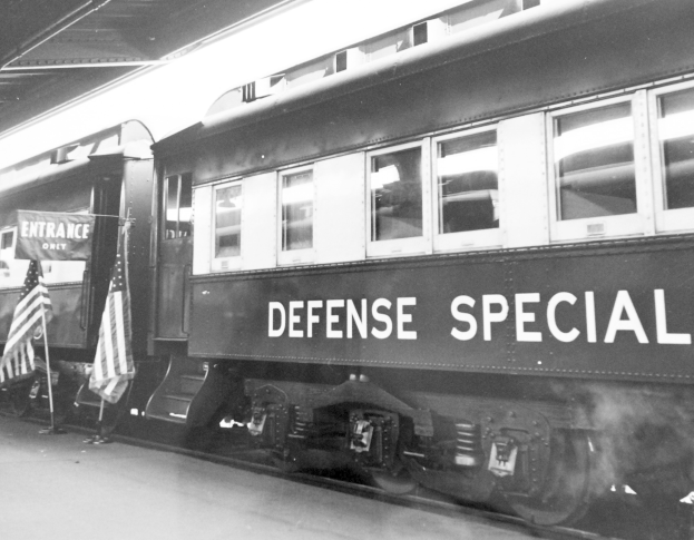 Schwarzes und weißes Foto eines Zuges mit der Aufschrift "Defense Special" auf Gleisen geparkt mit zwei Fahnen links und einer Decke oben.