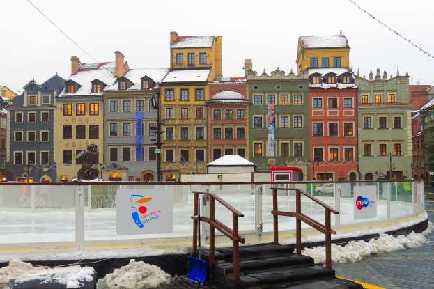 Eislaufbahn in einem Stadtplatz umgeben von Gebäuden, Treppen, Geländern, Informationsschildern, schneebedeckter Erde, Laternen, Bäumen und einem Himmel.