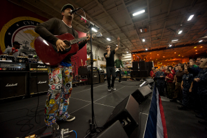 Chris Stapleton, der eine Mütze trägt, spielt Gitarre auf der Bühne vor einem Publikum, umgeben von einem Mikrofon, Lautsprechern, einer Flagge und anderen Instrumenten, mit einem Banner und Lichtern im Hintergrund.
