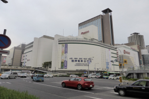 Eine belebte Stadtstraße mit Autos, hohen Gebäuden, Straßenlaternen, Schildern, Bäumen und einem klaren Himmel, mit einem Konzertsaal im Vordergrund.