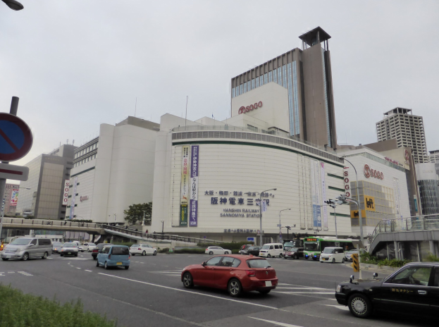 Eine belebte Stadtstraße mit Autos, hohen Gebäuden, Straßenlaternen, Schildern, Bäumen und einem klaren Himmel, mit einem Konzertsaal im Vordergrund.