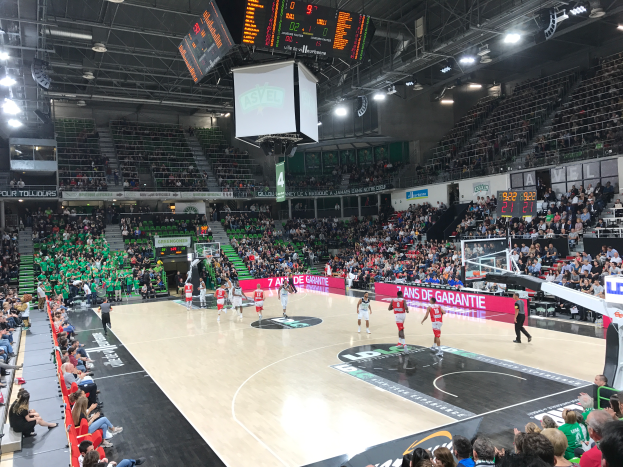 Ein Basketballspiel in einer großen Arena mit Zuschauern auf den Tribünen, Anzeigetafeln mit "Asvel vs Galatasaray" und einer beleuchteten Decke mit einem Bildschirm über dem Court.