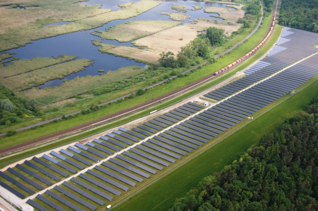 Luftaufnahme einer Solar-Farm mit Solar-Panelen auf einem Feld, umgeben von Bäumen, Gras, Pflanzen und Wasser, mit einem Zug auf einer nahen Bahnlinie.