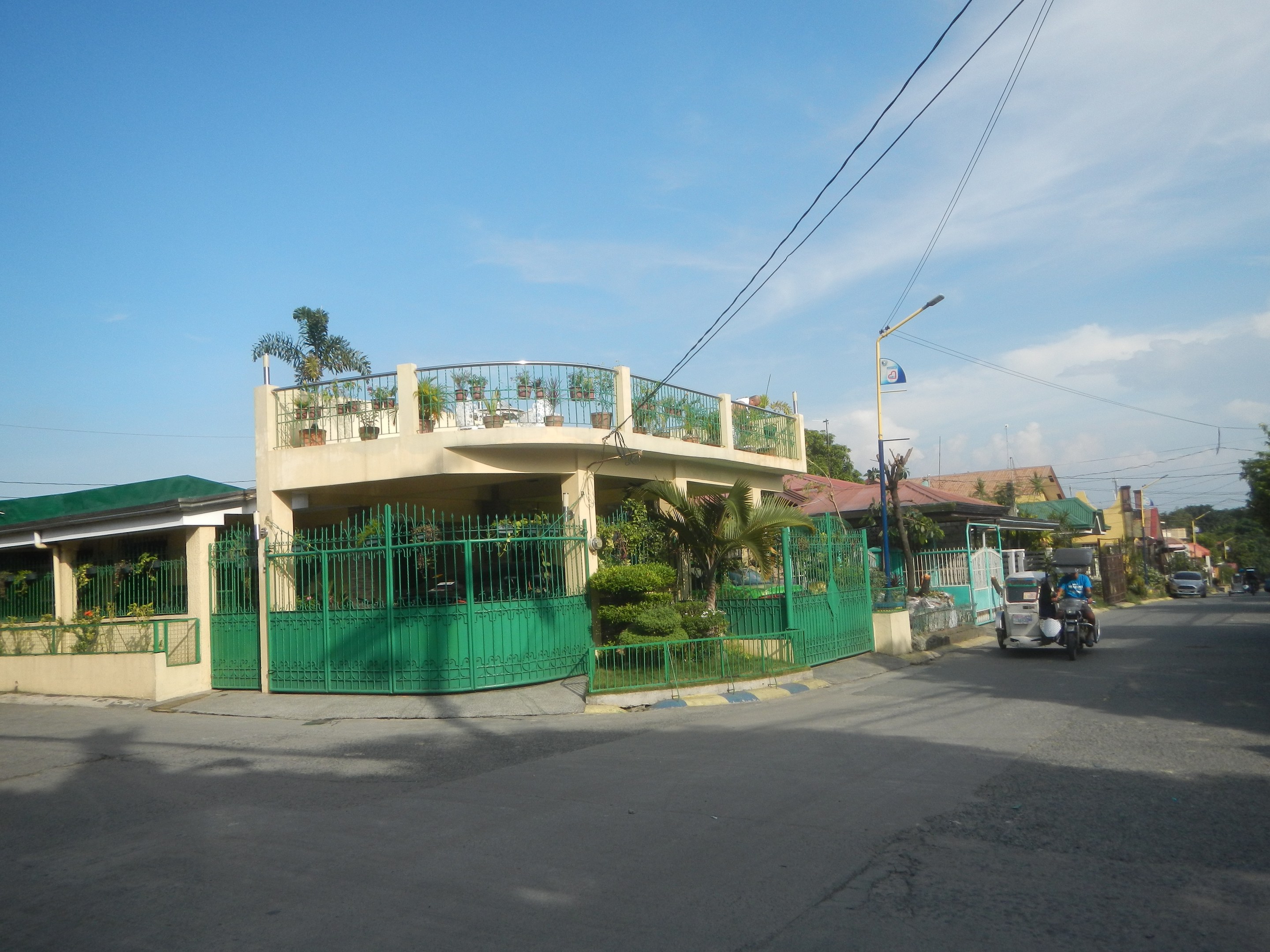 Eine Straße mit fahrenden Fahrzeugen, ein grünes Gebäude mit einem passenden Zaun, Geländern, Pflanzen, Bäumen, Strommasten mit Drähten und Lampen und eine Flagge, vor einem bewölkten Himmel.