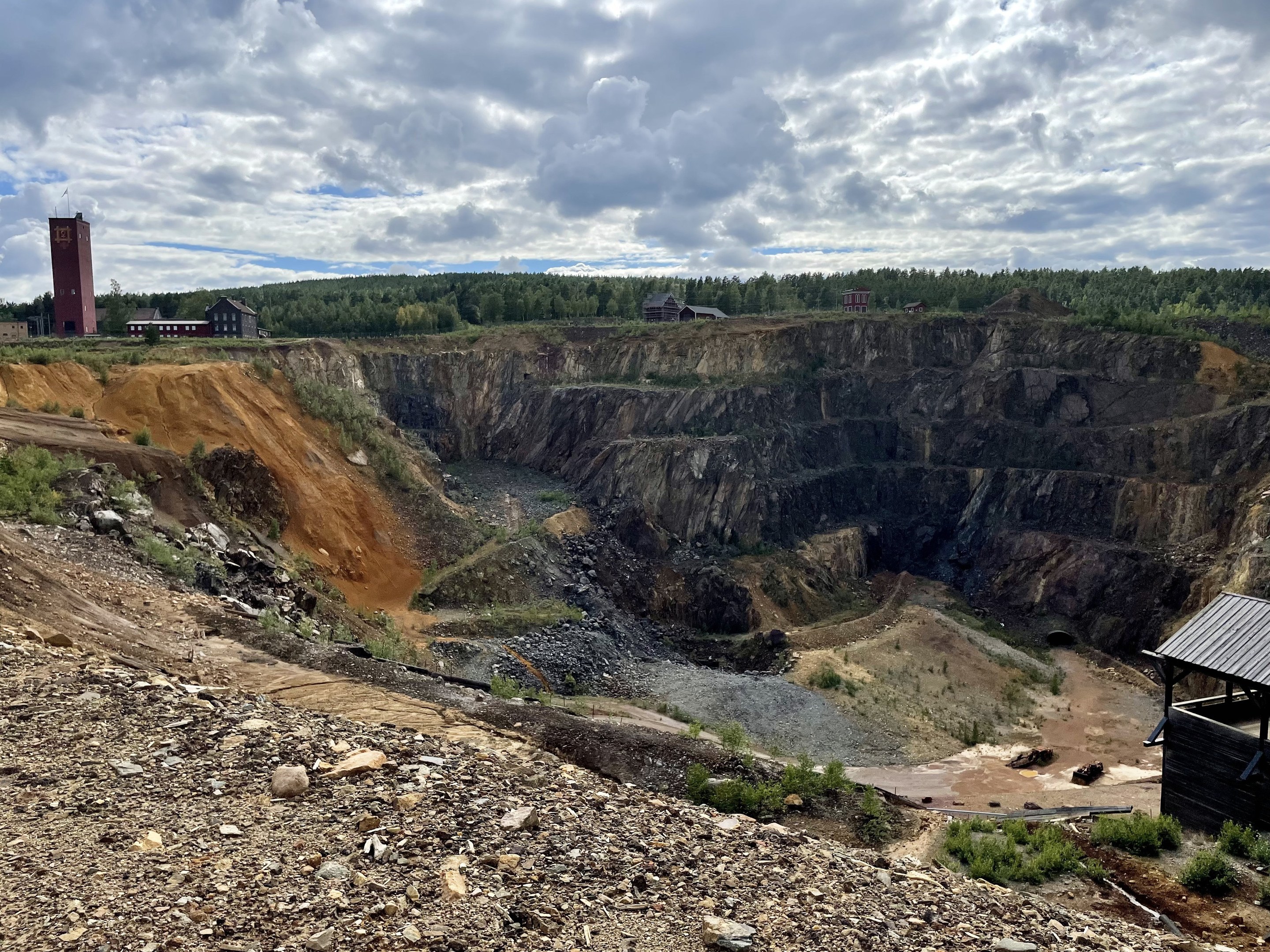 Großer Tagebau auf einem Schotterfeld umgeben von Bäumen, Gebäuden und einem Schuppen unter einem bewölkten Himmel, mit Steinen und Pflanzen auf dem Boden.