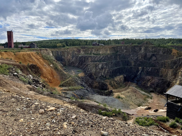 Großer Tagebau auf einem Schotterfeld umgeben von Bäumen, Gebäuden und einem Schuppen unter einem bewölkten Himmel, mit Steinen und Pflanzen auf dem Boden.