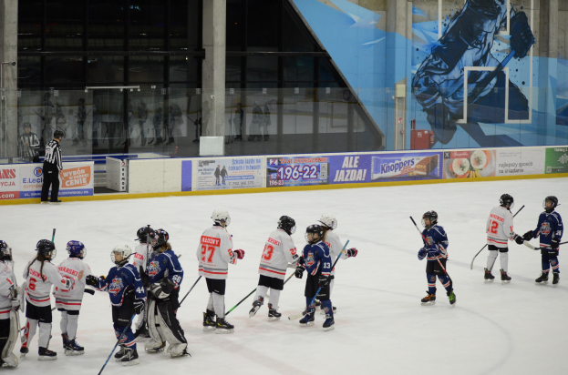 Gruppe junger Menschen, die Eis hockey auf einer Indoor-Eisbahn spielen, mit Helmen und Stöcken, einer Person auf der linken Seite und Bannern, Säulen und Glaswänden im Hintergrund.