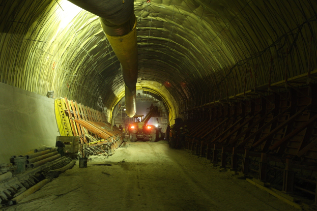 Baustelle mit einem großen mittigen Tunnel, Fahrzeugen, verstreuten hölzernen Gegenständen, Rohren und einer linken Wand, mit Beleuchtung von oben, die den Tunnel erhellt.