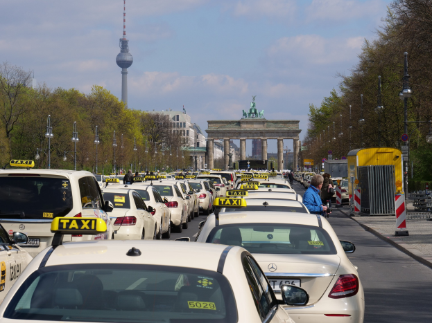 Eine belebte Straße in Berlin mit zahlreichen parkenden Taxis, Fußgängern auf dem Gehweg, gesäumt von Laternenpfählen, Bäumen und Gebäuden, mit einem fernen Bogen mit Statuen und einem Turm unter einem bewölkten Himmel.