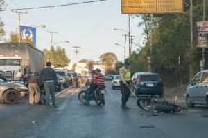 Gruppe von Menschen um ein verunglücktes Motorrad auf der Straßenseite mit mehreren Fahrzeugen, darunter ein Lastwagen, und Hintergrundelementen wie Bäumen, Pfosten, Lampen, Schildern und Himmel.