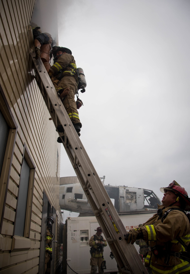 Feuerwehrleute mit Helmen klettern an einem Gebäude hoch, mit einem Fahrzeug und dem Himmel im Hintergrund.