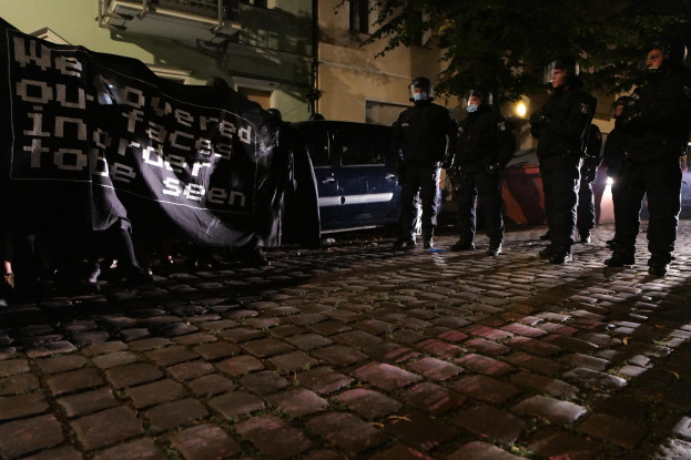 Gruppe von Polizisten in Helmen, die ein Banner auf einer Kopfsteinpflasterstraße bei Nacht halten, mit Fahrzeugen, Gebäuden, Bäumen und Straßenbeleuchtung im Hintergrund.