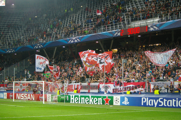 Ein Fußballspiel in einem hell erleuchteten Stadion mit einer großen Zuschauermenge, grünem Rasen, einem Torpfosten mit Bannern und einem Bildschirm in der linken oberen Ecke.
