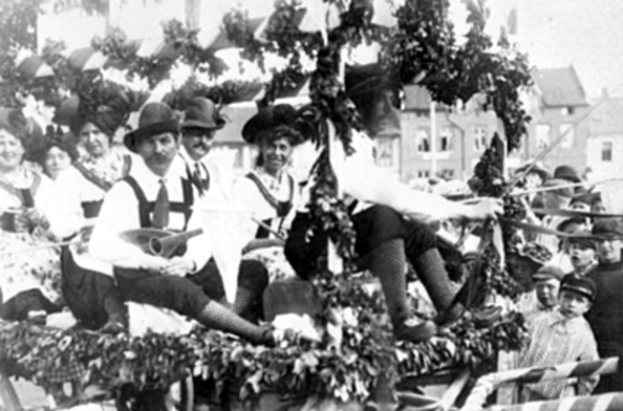 Schwarzes und weißes Bild von Menschen in traditioneller bayrischer Tracht auf einem geschmückten Wagen bei einem Umzug, einige spielen Instrumente, mit Gebäuden und einem klaren Himmel im Hintergrund.